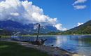 Ein ruhiger See umgeben von Bergen und grünen Wiesen. Die Wolken sind groß und weiß vor einem klaren blauen Himmel.