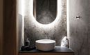 A modern bathroom with elegant lighting and a round mirror. A minimalist wooden vanity top with a white basin and fluffy slippers underneath.