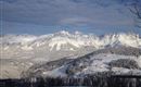 Eine winterliche Berglandschaft mit schneebedeckten Gipfeln und Wäldern. Der Himmel ist klar und beleuchtet.