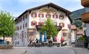 A historic building with colorful windows and a balcony. Two cyclists ride past a shady tree.
