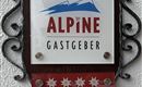 Ein Logo mit dem Schriftzug "ALPINE GASTGEBER" und stilisierten Bergen. Die Farben sind rot, blau und weiß, mit Edelweiß-Blumen am unteren Rand.