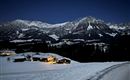 Eine verschneite Landschaft bei Nacht mit beleuchteten Häusern. Im Hintergrund erheben sich beeindruckende Berge unter einem klaren Sternenhimmel.