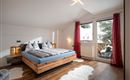 Ein gemütliches Schlafzimmer mit einem großen Doppelbett und modernen Holzdetails. Große Fenster lassen viel Tageslicht herein und bieten Zugang zu einem Balkon.
