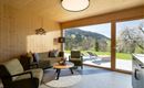 A modern living room with wooden walls and large windows. The view looks out over a green landscape with mountains in the background.