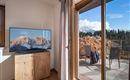 A cozy room with a television and a view of the mountains. Outside is a terrace with chairs and a beautiful landscape.