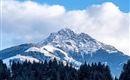 A majestic mountain with a snow-capped peak and green forests in the foreground. The sky is clear with a few clouds.