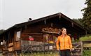 Een vrouw in een oranje jas wandelt op een weiland. Op de achtergrond staat een houten huis met bloembakken.