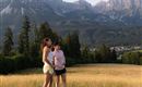 Twee personen staan op een groene weide met uitzicht op majestueuze bergen op de achtergrond. Het is een zonnige dag en het landschap straalt een rustige sfeer uit.