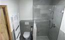 Een moderne badkamer met een glasdouche en grijze tegels. Rechts bevindt zich een toilet en een handdoekradiator.