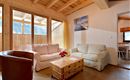 Ein gemütliches Wohnzimmer mit Holzdecke und großen Fenstern. Es gibt bequeme Sofas und einen Holztisch mit einem Blumenarrangement.