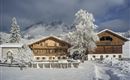 A picturesque winter landscape with snow-covered cottages and trees. In the background, majestic mountains rise under a clear sky.