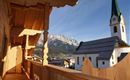 Ein Balkon mit Holzverkleidung und Blick auf eine malerische Kirch und majestätische Berge. Der Himmel ist klar und die Architektur ist traditionell.