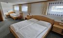 A simple guest room with two beds and a large window. The walls are bright and there is wooden furniture.