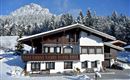 A beautiful chalet in the snow with a picturesque mountain backdrop. The surroundings are filled with many fir trees.