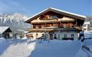 A traditional wooden house in the snow with mountains in the background. It is a clear, sunny day.