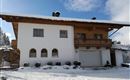 A cozy house in alpine style with a covered balcony. The surroundings are snow-covered and the sky is partly cloudy.