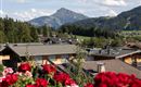 Eine malerische Berglandschaft mit einfachen Häusern und üppigem Grün. Im Vordergrund blühen rote Blumen. Ausblick auf das Kitzbühler Horn vom Balkon aus.