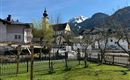 An idyllic village with beautiful houses and a church tower. In the background, snow-covered mountains and a clear blue sky can be seen.