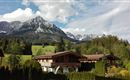 Eine idyllische Berglandschaft mit schneebedeckten Gipfeln und sattgrünen Wiesen. Im Vordergrund steht ein gemütliches Holzhaus umgeben von Bäumen.