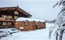 A traditional wooden house with a snow-covered roof sits in a wintry landscape. Snow blankets the ground and the trees all around.