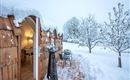 A cozy wooden house in the snow with a snowy landscape. The entrance is inviting, with a table and chairs outside.
