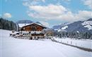 A picturesque mountain landscape in winter with snow-covered fields. In the foreground, there is a traditional wooden house against an impressive mountain backdrop.