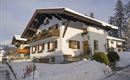 A traditional house in the snow with a steep roof and wooden balconies.  
Surrounded by a winter landscape and tall trees.
