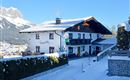 A picturesque house in the snow with a beautiful mountain landscape in the background. The sky is clear and blue, creating a tranquil winter atmosphere.