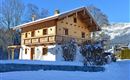 Ein traditionelles Holzhaus in einer schneebedeckten Landschaft. Im Hintergrund sind Bäume und Berge zu sehen.