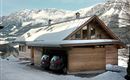 A modern wooden house in the snow, surrounded by mountains. Two cars are parked in the garage.