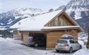 A cozy wooden house in the snow with a garage area. Impressive mountains can be seen in the background.