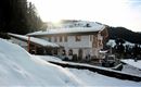 A charming chalet in the snow with mountains in the background. The sun gently shines on the building and the surroundings.