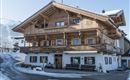 A traditional wooden house with several balconies and large windows. It stands in a snow-covered landscape and seems to be nestled in a picturesque mountain setting.