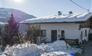 A cozy house in the snow with a snow-covered roof. In the background, there are mountains and a clear blue sky.