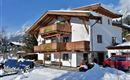 A beautiful old house with wooden balustrades, surrounded by snow. In the background, mountains and blue sky can be seen.