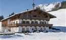 A traditional alpine house in the snow, surrounded by a winter landscape. The mountains are visible in the background and the sky is clear.