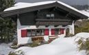 A charming Alpine house with red shutters, surrounded by snow. The winter magic is enhanced by the snowy landscape and the fir trees.