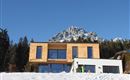 A modern house made of wood and concrete, surrounded by snow-covered mountains. The clear blue sky gives the scene a calm atmosphere.
