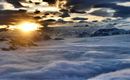 A sunrise over snow-covered mountains. The sky is filled with dramatic clouds and a sea of mist.