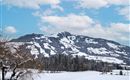 Eine verschneite Landschaft mit einem Berg im Hintergrund. Der Himmel ist blau und hat einige Wolken.
