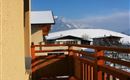 A balcony with a wooden railing that offers a picturesque view of snow-covered mountains. The sky is clear and blue, which impressively highlights the winter landscape.