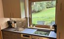 Eine moderne Küche mit einer großen Fensterfront, die einen Blick auf den Garten bietet. Der Raum ist hell und gut ausgestattet mit Kochgeräten.