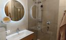 Een moderne badkamer met een glazen douche en een houten wastafel. Een ronde spiegel met verlichting is aan de muur bevestigd.