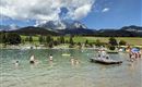 A clear lake surrounded by green meadows and mountains in the background. People are enjoying the water and the sun.