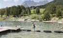 A boy is jumping on foam islands in the water. In the background, swimmers and a green meadow with mountains can be seen.