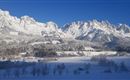An impressive winter landscape with snow-covered mountains and a clear blue sky. In the foreground, a quiet, snowy plain stretches out.