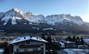 A picturesque mountain panorama with snow-capped peaks and a clear sky. In the foreground, a few houses and trees can be seen.