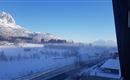 A winter landscape with snow-covered mountains and a clear blue sky. In the foreground, there is a road and some buildings visible.