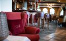 Ein gemütlicher Sessel mit einem Kissen in rot steht in einem stilvollen Restaurant. Im Hintergrund sind elegante Stühle und eine Bar sichtbar.