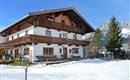 Ein traditionelles Haus im alpine Stil, umgeben von Schnee. Im Hintergrund sind Berge und ein blauer Himmel zu sehen.